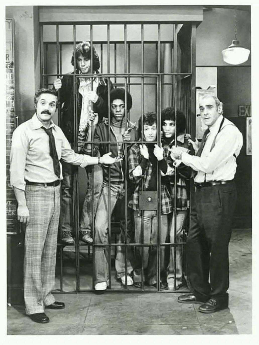 A black and white promotional photo from the 1970s TV show 'Welcome Back, Kotter', featuring the main cast. The 'Sweathogs' (including a young John Travolta) are depicted inside a jail cell, while two adult characters, likely teachers or authority figures, stand outside. The scene suggests a comedic or lighthearted moment, typical of the show's pre