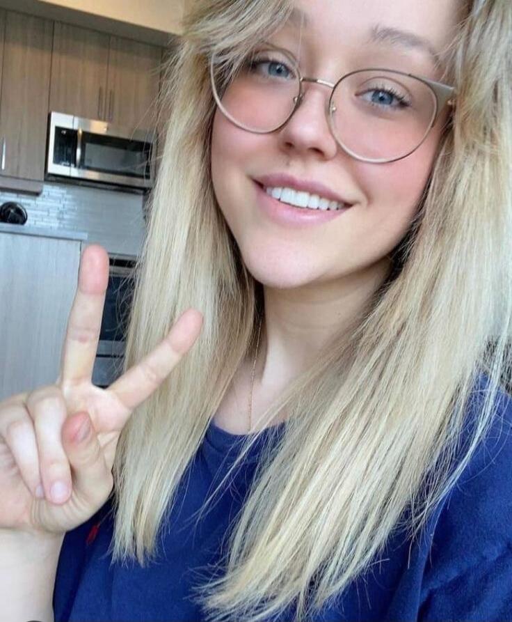 A young blonde woman with glasses is smiling and making a peace sign. She is wearing a blue t-shirt. The background shows a kitchen.