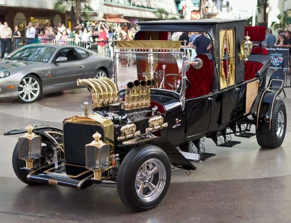 A highly customized black and gold vintage-style hot rod car with red velvet interior and prominent gold exhaust pipes, parked on a street or plaza with people and another car in the background.