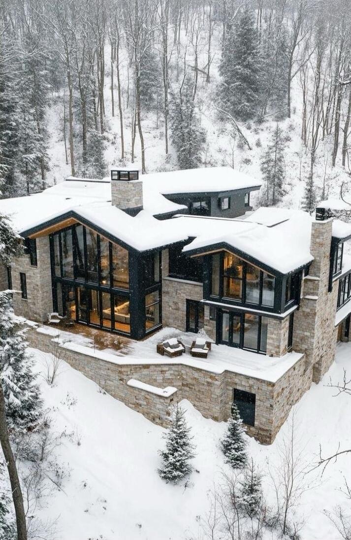 A large modern stone house in a snowy forest setting, featuring extensive glass walls and multiple interconnected wings. Snow covers the roof and surrounding trees.