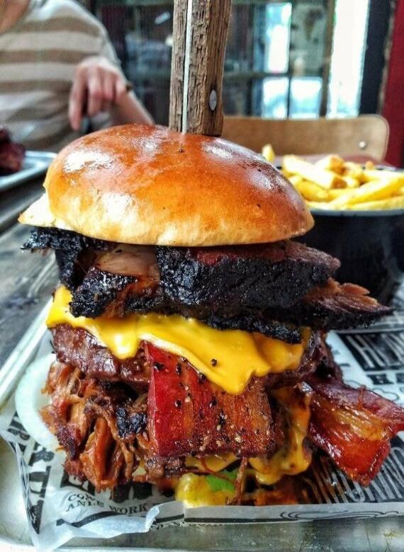 A large, multi-patty cheeseburger with bacon and melted cheese stacked high, served on a glossy bun with fries in the background.
