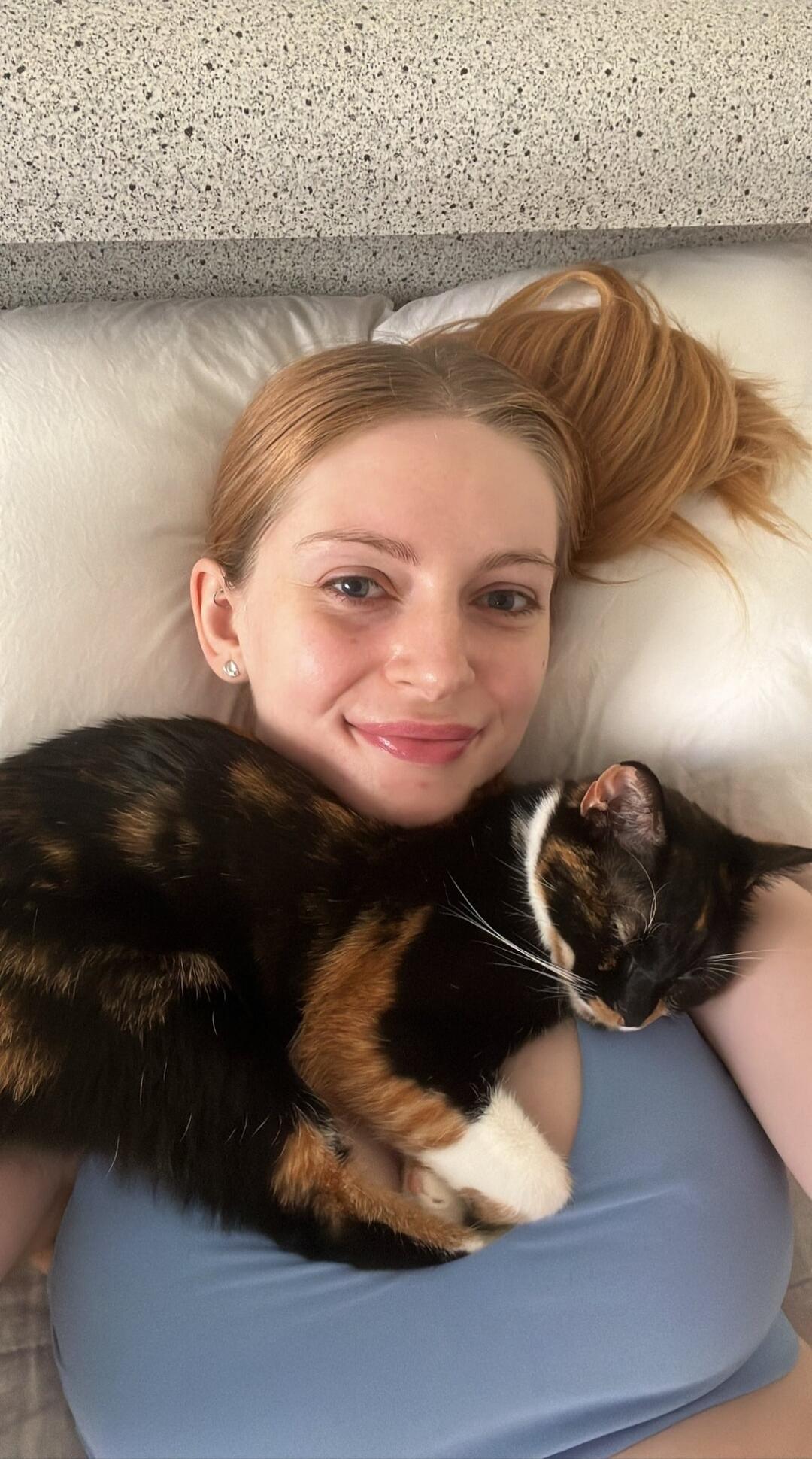 A woman with red hair is lying in bed, smiling at the camera, with a calico cat sleeping peacefully on her chest.