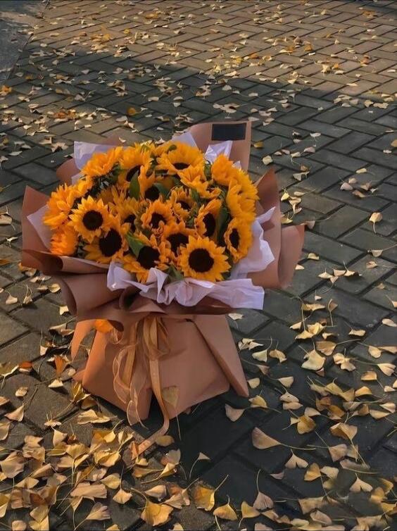 A bouquet of bright sunflowers wrapped in brown paper on a brick pathway with scattered autumn leaves.