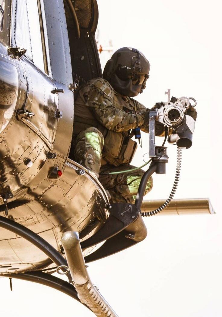 A soldier in camouflage gear operating a mounted machine gun from a helicopter.