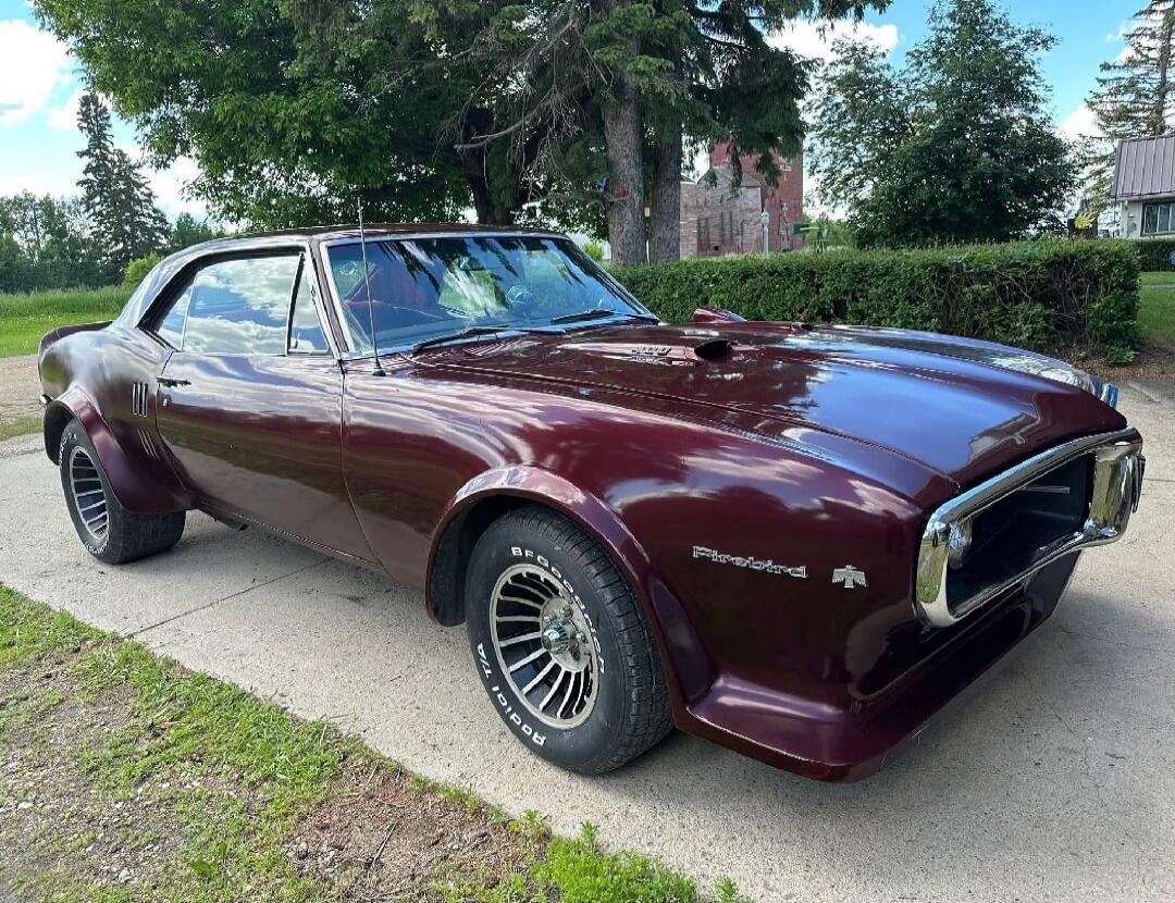 A classic maroon Pontiac Firebird car is parked on a driveway. The car has wide fenders and custom wheels. The 'Firebird' emblem is visible on the front fender.