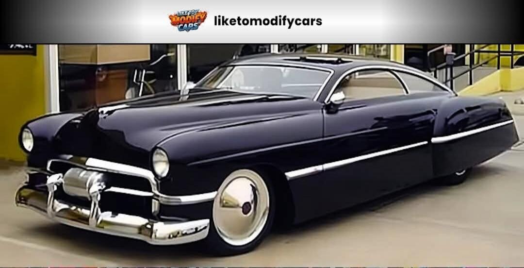 LIKETOMODIFYCARS liketomodifycars. A custom dark-colored vintage car, possibly a Cadillac, with a sleek design, chopped top, and custom wheels, parked outdoors.