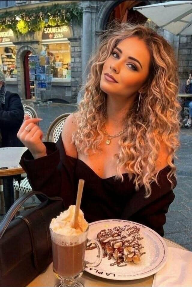 A young woman with curly blonde hair is sitting at an outdoor cafe table. She is wearing an off-the-shoulder top, revealing her shoulders and décolletage. On the table, there is a glass of hot chocolate with whipped cream and a plate with waffles or pancakes topped with chocolate and nuts. In the background, a shop sign reads 