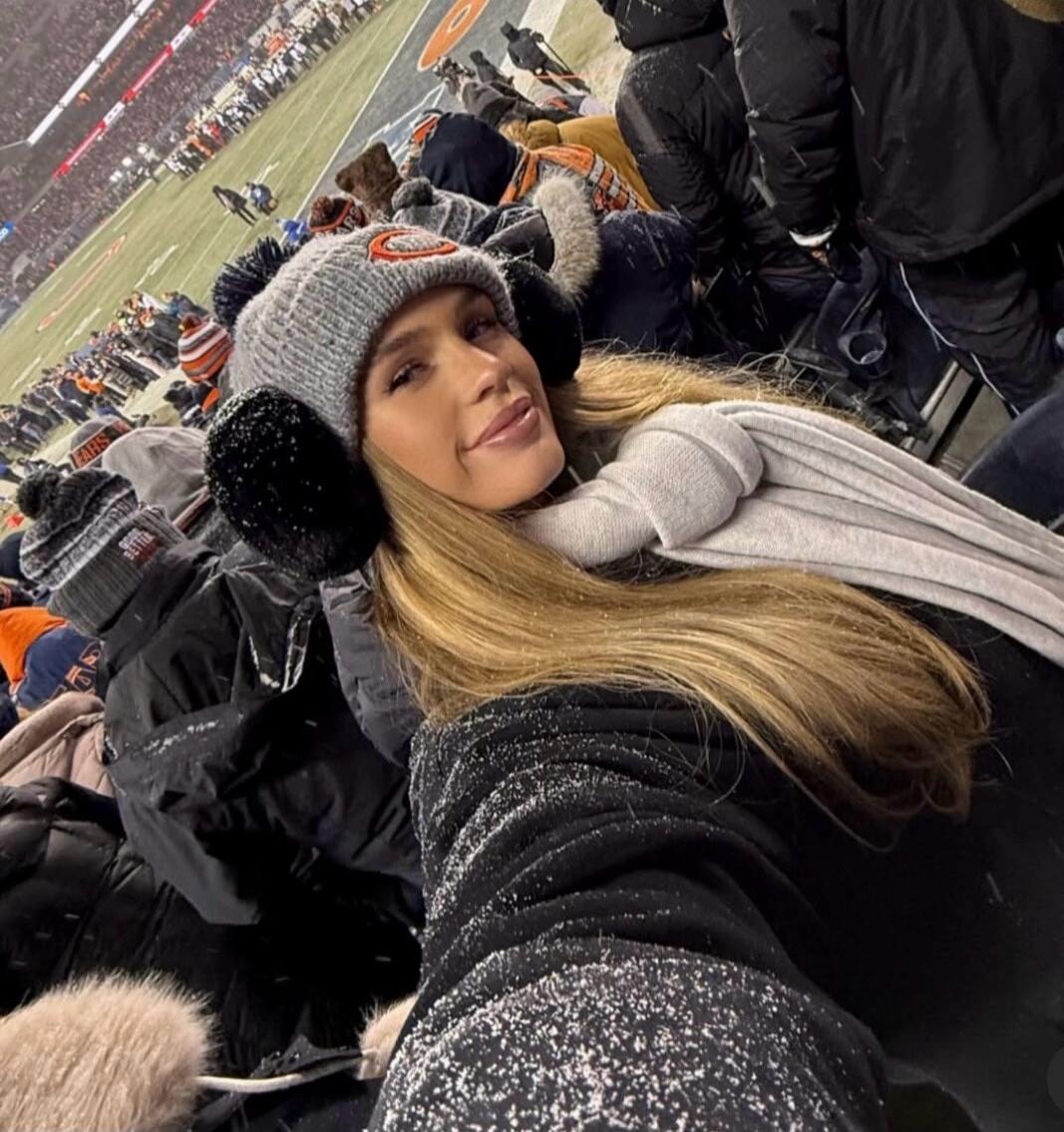 A woman taking a selfie in a snowy stadium, wearing a grey beanie with black pom-poms and a scarf, with fans in the background.