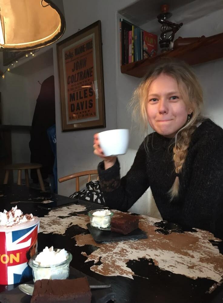A young woman with blonde hair is sitting at a table, holding a white cup. On the table, there are brownies, whipped cream, and a mug with a Union Jack flag design that says 'ON'. In the background, a framed poster features the names 'JOHN COLTRANE' and 'MILES DAVIS'.
