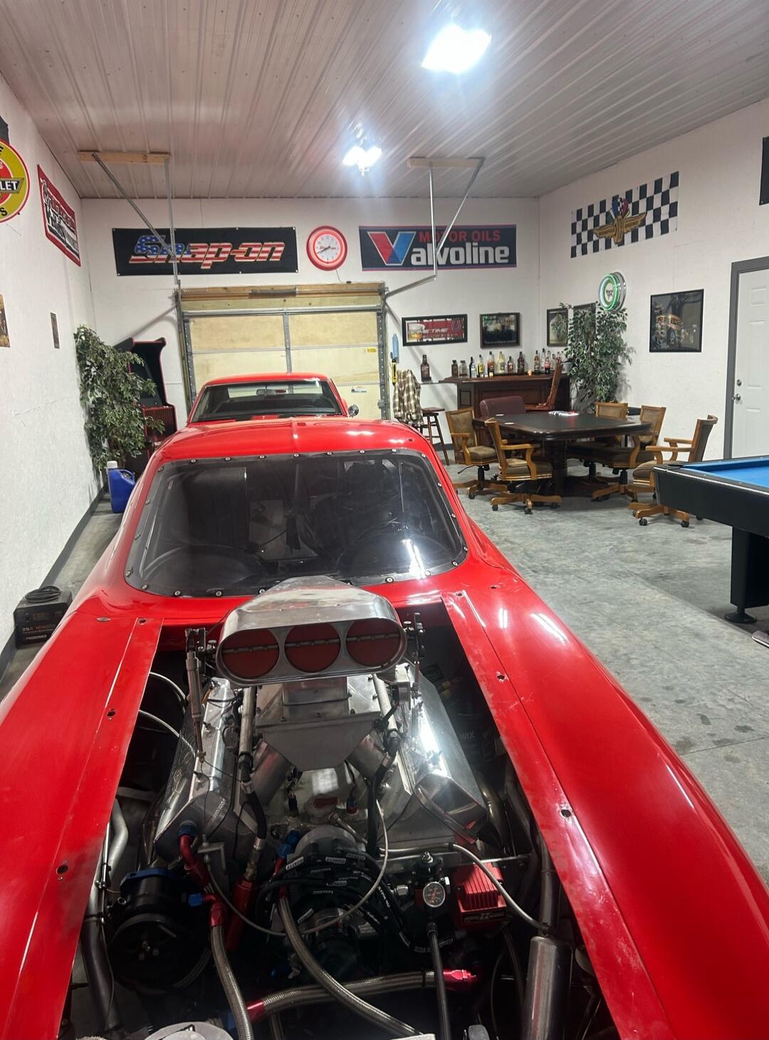 A red drag racing car with a powerful engine and supercharger is parked in a spacious garage. The garage is decorated with automotive signs like Snap-on, Valvoline, and Chevrolet, and features a recreational area with a bar, poker table, and pool table. Another car is visible in the background.