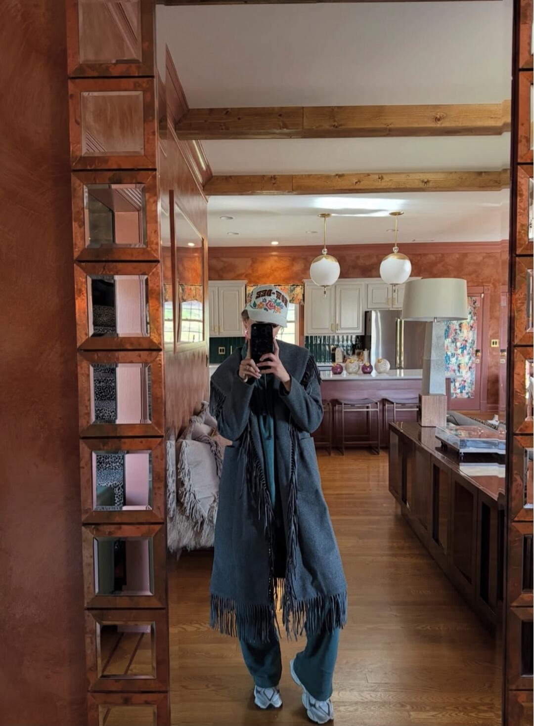 Person taking a mirror selfie in a spacious, wood-paneled kitchen area. The individual is wearing a long denim poncho with fringes, jeans, sneakers, and a bucket hat, standing in front of a large wooden-framed mirror.