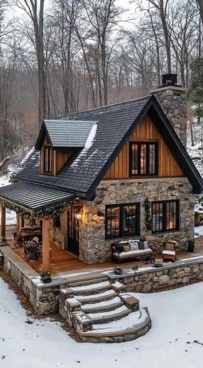 A stone cottage with a steep slate roof, wooden accents, string lights, and a snow-covered porch in a wintry forest.