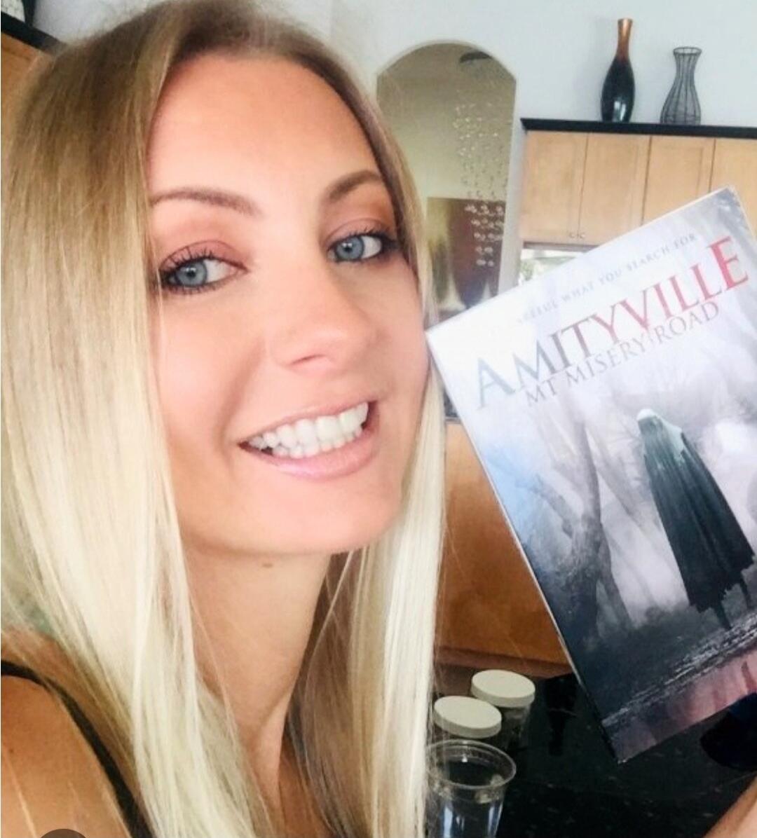 A woman is smiling and holding a DVD case for the movie 'Amityville Mt Misery Road'. The text on the case also says 'See what you search for'.