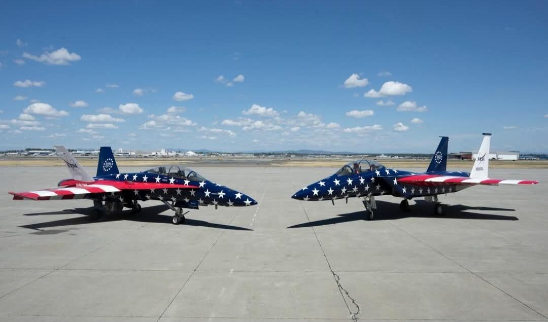 Two NASA F-15 Eagle fighter jets, painted in a patriotic American flag scheme with blue, white stars, and red and white stripes, are parked on an airfield under a clear blue sky with scattered clouds. The jets are facing each other, showcasing their distinctive livery.