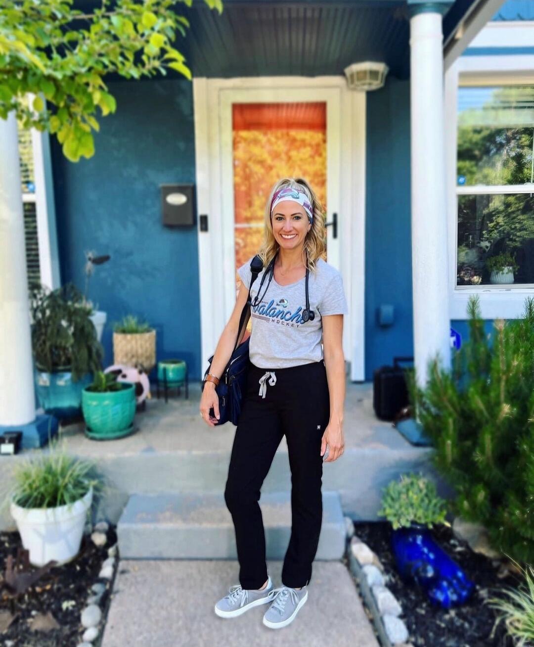 A smiling woman standing on a front porch in casual clothes (gray t-shirt, black joggers, gray sneakers) with a headband and a bag.
