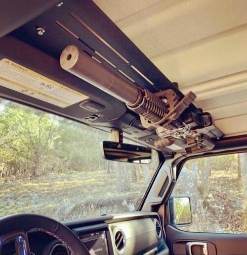 A warning label is partially visible. A rifle with a suppressor is mounted on the ceiling of a car, likely a Jeep, in an outdoor setting.