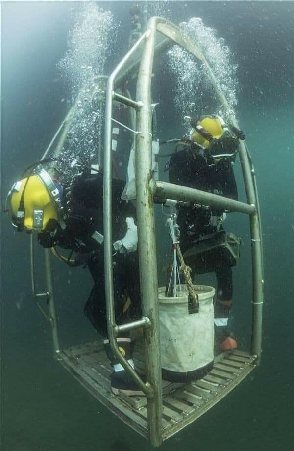 Two divers in a metal underwater cage with bubbles rising around them; a bucket hangs inside the cage.