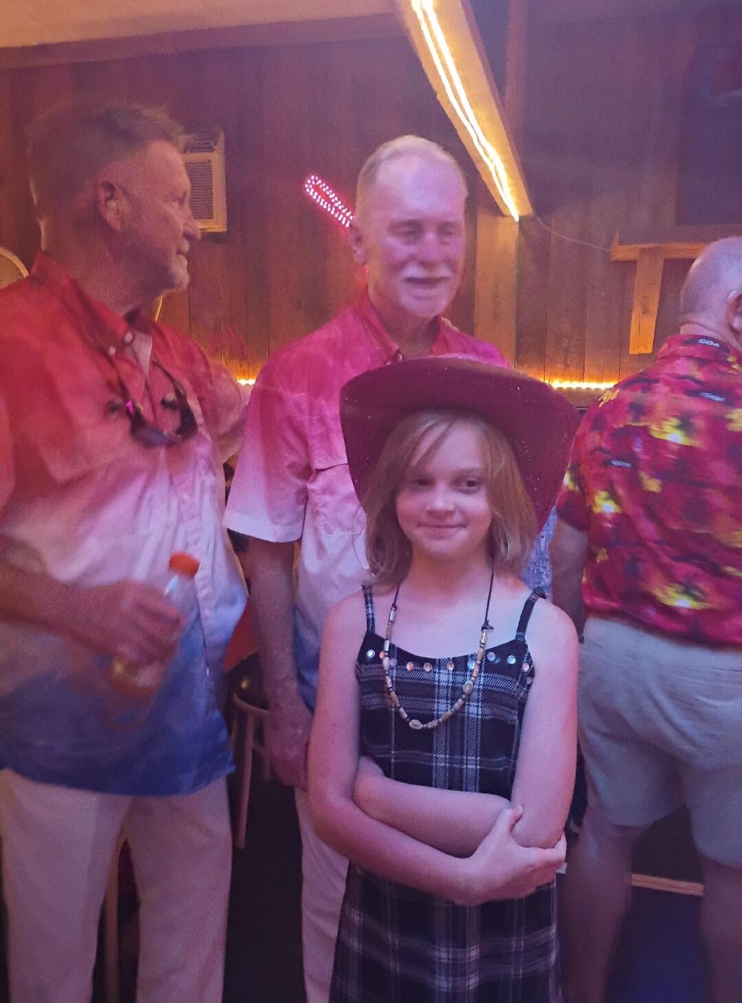 A young girl wearing a cowboy hat and a plaid dress stands in front of two adult men at an indoor gathering. The men are casually dressed, one in a red, white, and blue shirt, the other in a floral shirt. The atmosphere is warm and friendly.