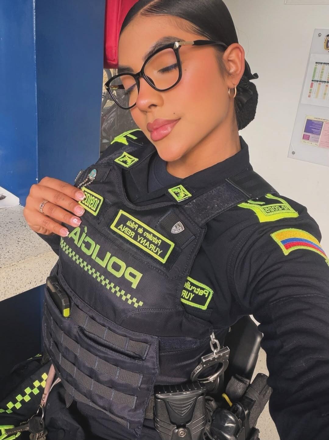 A woman in a Colombian police uniform, wearing glasses, with her eyes closed and a gentle smile. Text on uniform includes 'POLICIA' and a Colombian flag patch.