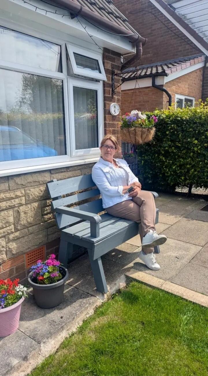 A woman is sitting on a grey wooden bench outside a brick house. She is wearing a white jacket, light brown pants, and white sneakers. She has glasses and is holding a phone. There are potted flowers on the ground and a hanging basket with flowers near the house. The house number '85' is visible on the wall.