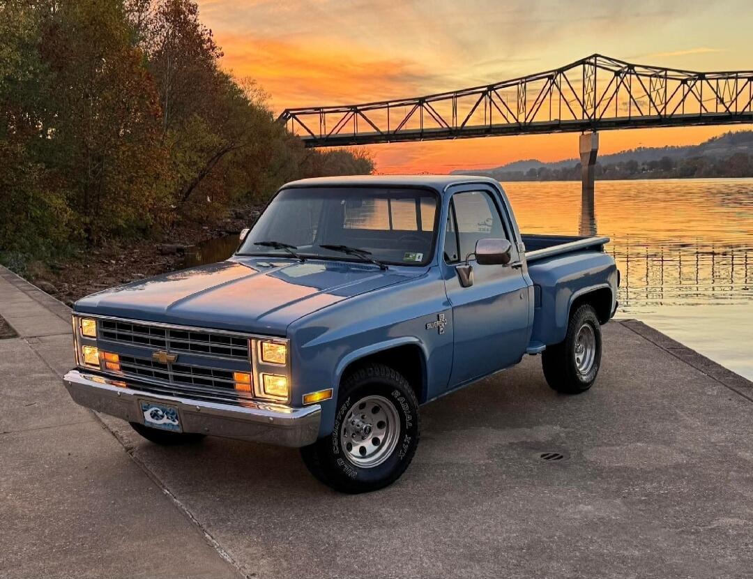 A vintage blue pickup truck parked on a concrete riverfront at sunset, with a bridge spanning the river in the background.