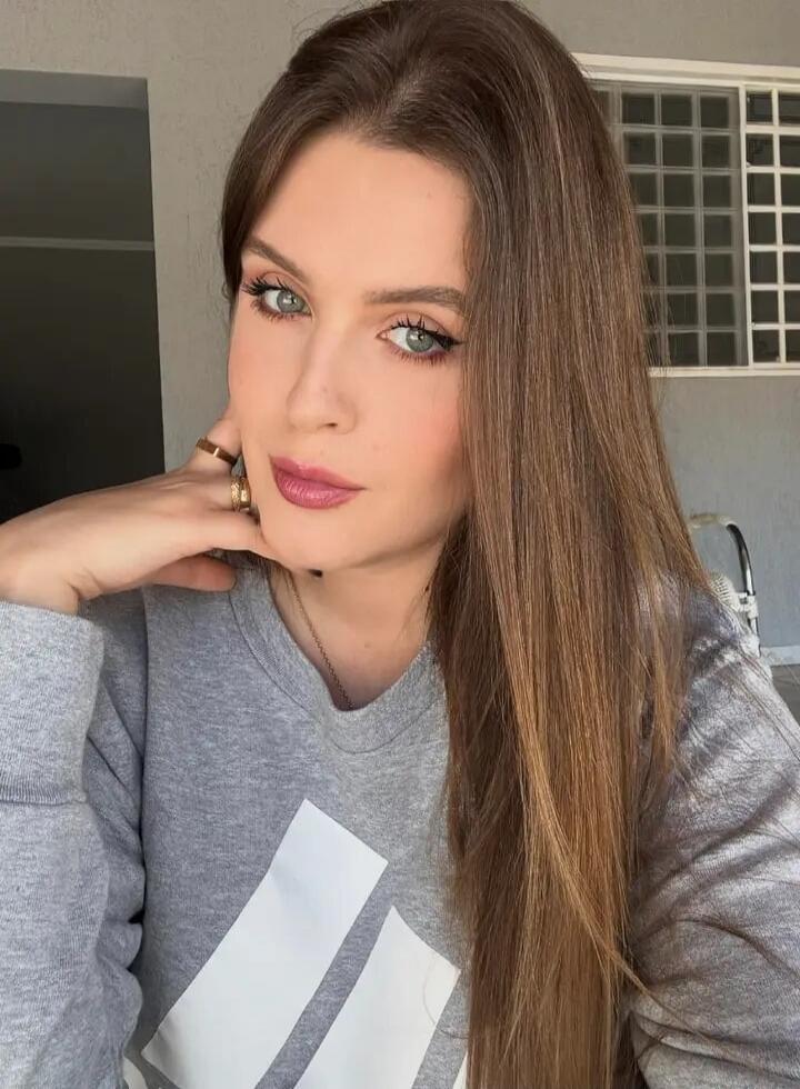 A woman with long brown hair and green eyes, wearing a grey sweatshirt with an Adidas logo, looking at the camera. Her hand is near her chin, showing rings.