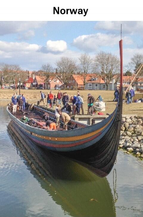 Norway. A large, traditional wooden boat, resembling a Viking ship, is docked in calm water. Several people are gathered on and around the boat, appearing to be engaged in maintenance or preparation activities. In the background, there are houses and trees under a partly cloudy sky.