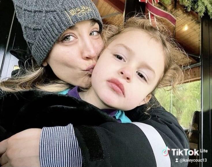 A woman in a grey beanie is kissing a child on the cheek. The child looks serious. The woman is smiling slightly. The TikTok Lite watermark with username @kaleycouc13 is visible.