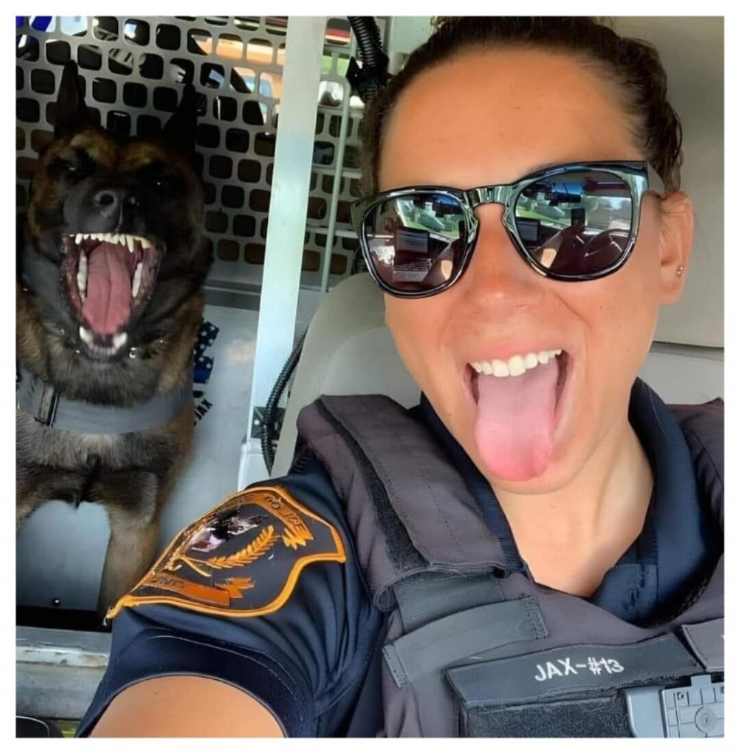 A female police officer in uniform and sunglasses takes a selfie with her K9 partner. The officer is sticking her tongue out playfully, while the police dog in the background has its mouth wide open, showing teeth, mimicking the officer's playful expression. Text on uniform: 'POLICE', 'JAX-#13'. Text on dog's harness: 'JAX'.