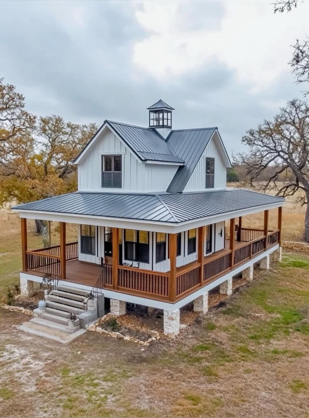 A two-story white farmhouse with a wraparound porch on a raised foundation, metal roof, and a cupola, set in a rural landscape.