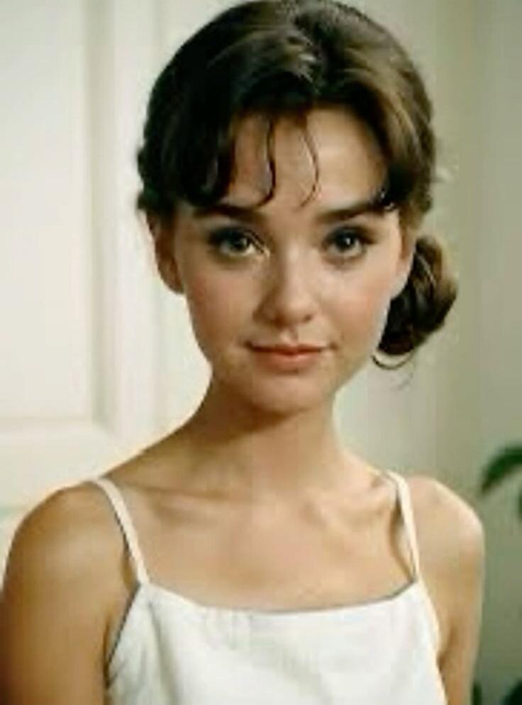 A young woman wearing a white sleeveless top, posing indoors.