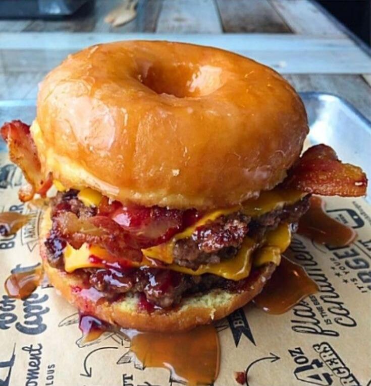 A burger made with glazed donuts as buns, featuring two beef patties, melted cheese, and crispy bacon. Sauce is dripping from the burger onto the paper wrapper. Visible text on wrapper includes 
