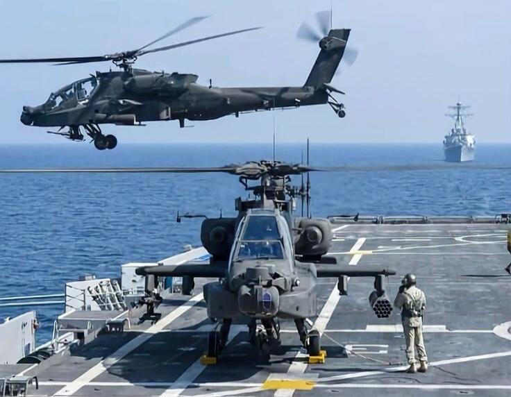 Two military helicopters, likely Apache, are seen on and above the deck of a warship at sea. One helicopter is on the deck, while another is in the air, either taking off or landing. A person in military uniform stands near the helicopter on the deck. Another warship is visible in the distance.