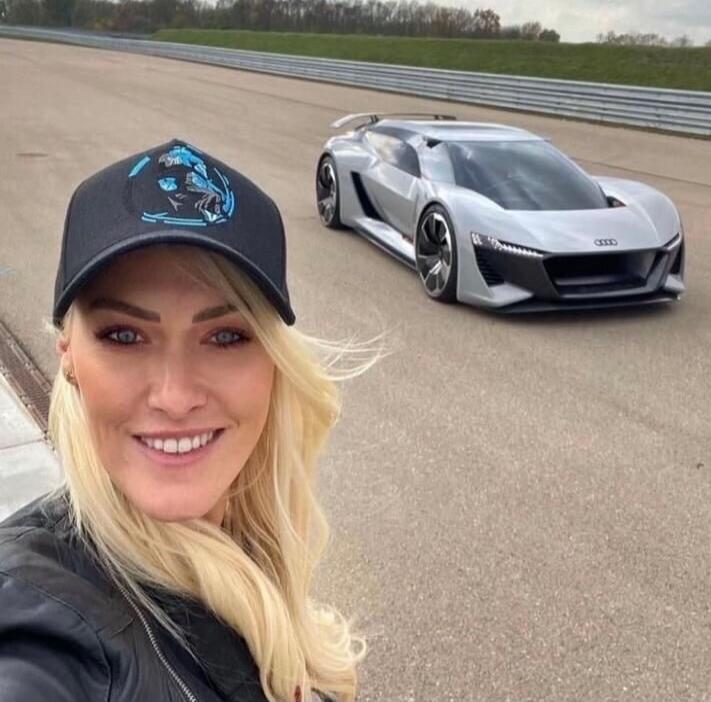 A woman takes a selfie with a silver futuristic sports car on a race track. The car appears to be a high-end concept or supercar with a low profile and sharp angles. The woman is wearing a dark cap and jacket, smiling at the camera.