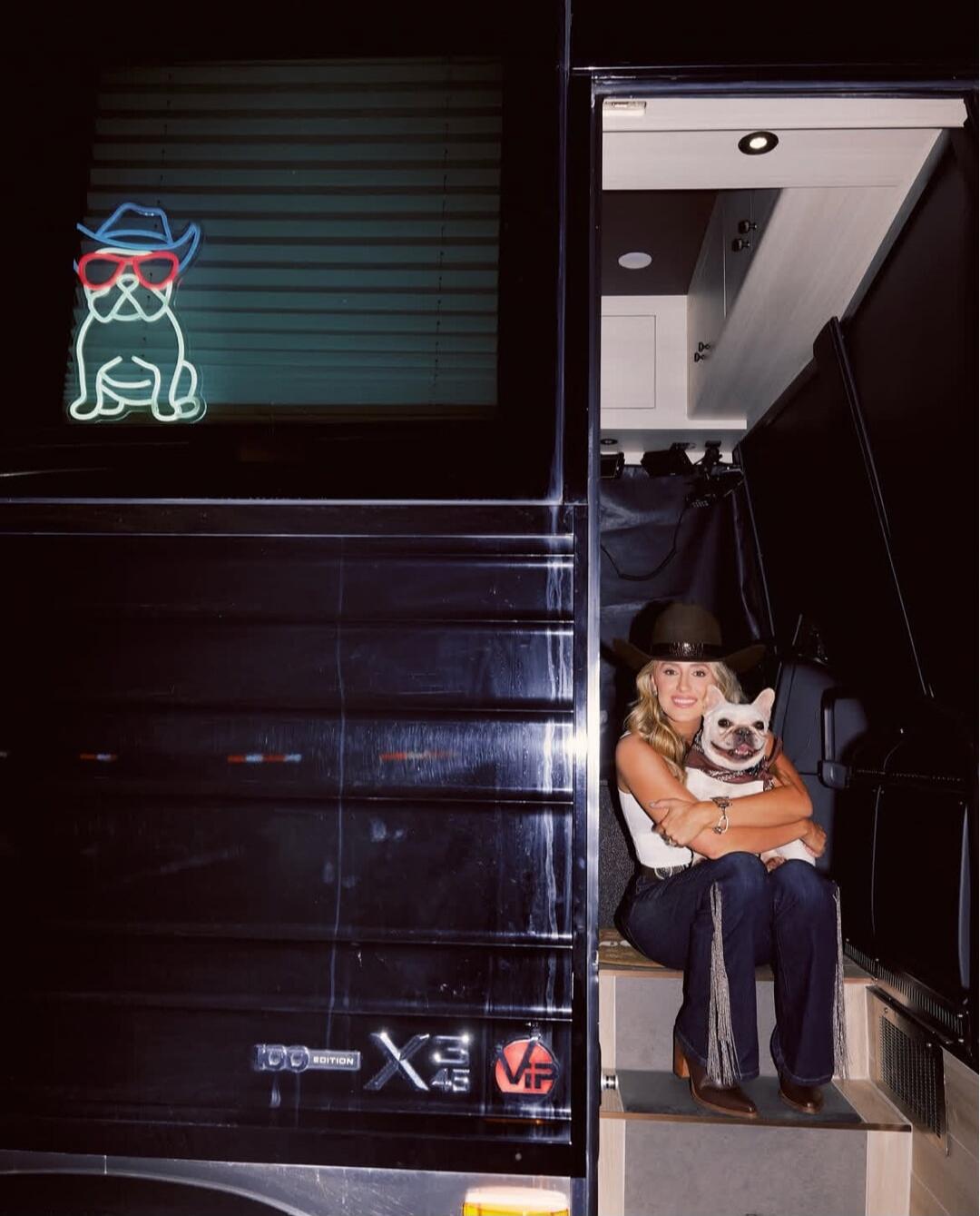 100 EDITION X3 45 VIP. A neon sign of a dog wearing a cowboy hat and sunglasses is visible on the window. A woman in a cowboy hat and fringed jeans is sitting on the steps of a vehicle, holding a white French Bulldog.