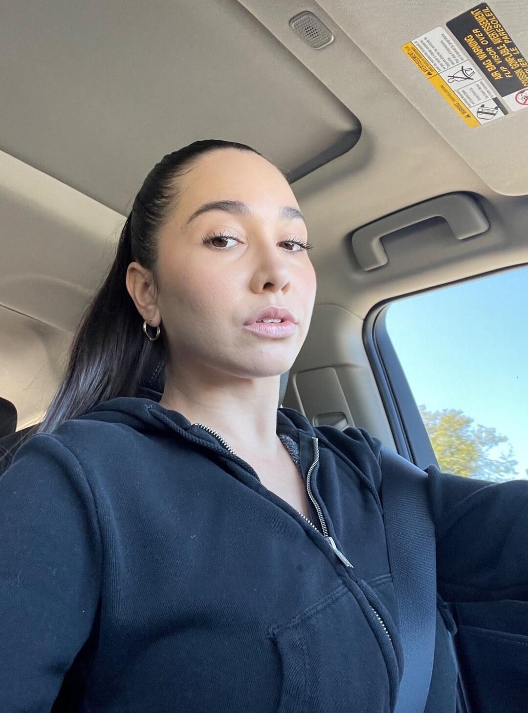 A woman with dark hair pulled back, wearing a black zipped hoodie, is sitting in a car and looking directly at the camera. On the car's sun visor, a warning label is visible, which includes the text 'AIR BAG WARNING' and 'COUSSIN GONFLABLE AVERTISSEMENT'.