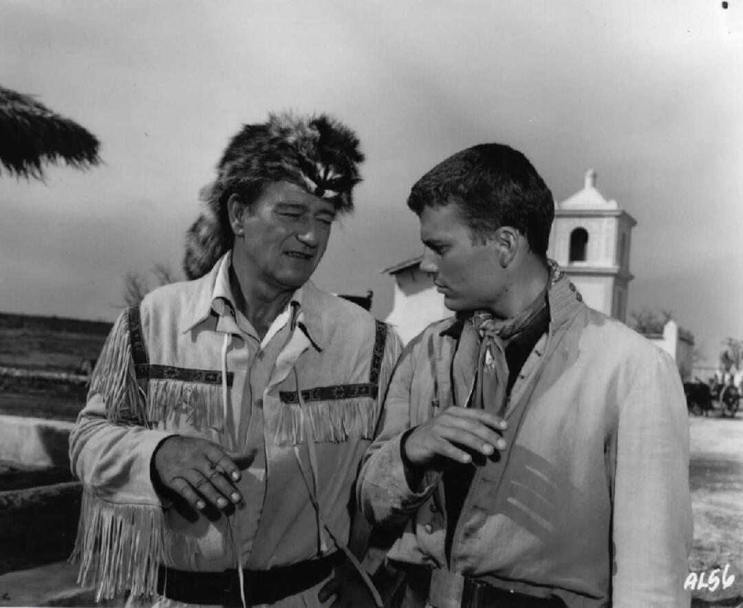 AL56. Two men, one in a coonskin cap and fringed jacket, the other in a simple shirt, are engaged in conversation. A mission-style building is visible in the background.