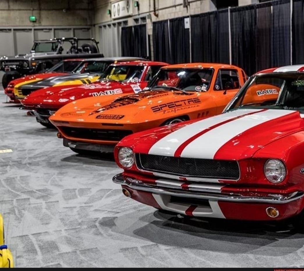 A line of classic American muscle cars at an indoor car show. The cars include a red Ford Mustang with white racing stripes, an orange Chevrolet Corvette with 'SPECTRE PERFORMANCE' and 'BAER' branding, and other vintage vehicles. 'CROWER' is also visible on one of the cars.