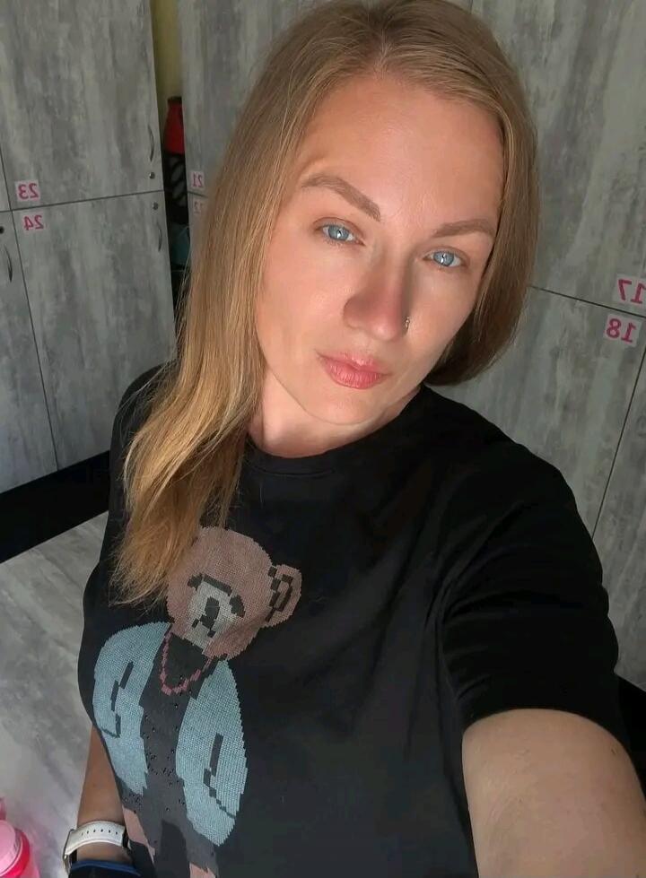 A woman with blonde hair and blue eyes takes a selfie in what appears to be a locker room. She is wearing a black t-shirt with a pixelated teddy bear design. Lockers are visible in the background.