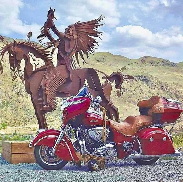 A red Indian motorcycle parked in front of a large metal sculpture depicting a Native American warrior on horseback, set against a backdrop of rolling hills and a cloudy sky.