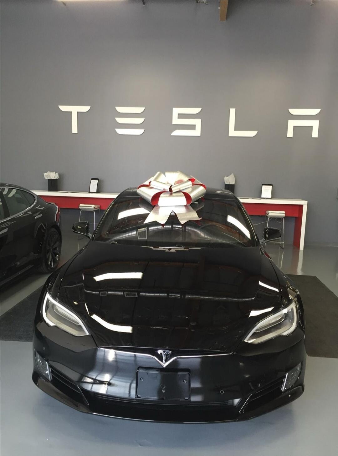 Tesla logo on wall; black Tesla car in a showroom with a large white and red bow on the roof.