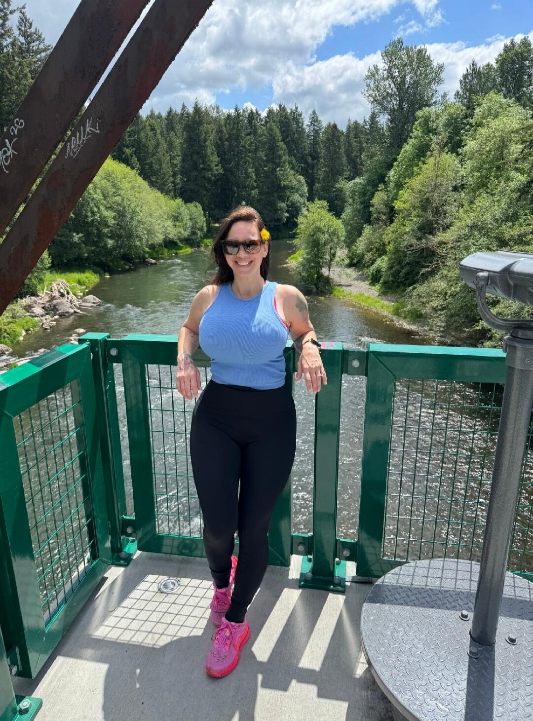 A woman stands on a bridge, smiling at the camera. She is wearing a light blue ribbed tank top, black leggings, and bright pink athletic shoes. She has sunglasses on and a yellow flower tucked behind her ear. In the background, a river flows through a lush green forest under a partly cloudy sky. There is some graffiti on the metal structure of the 