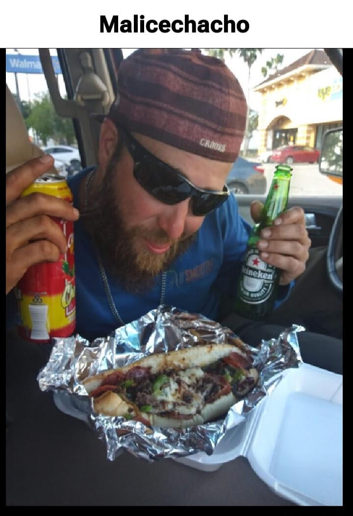 Malicechacho. A man with a beard and sunglasses is in a car, holding a red can in his left hand and a Heineken beer bottle in his right hand. He is looking down at a large sandwich, possibly a cheesesteak, wrapped in foil and placed in a styrofoam container. The text 
