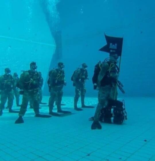 A group of divers in full gear are seen underwater in what appears to be a swimming pool. One diver is holding a black flag.