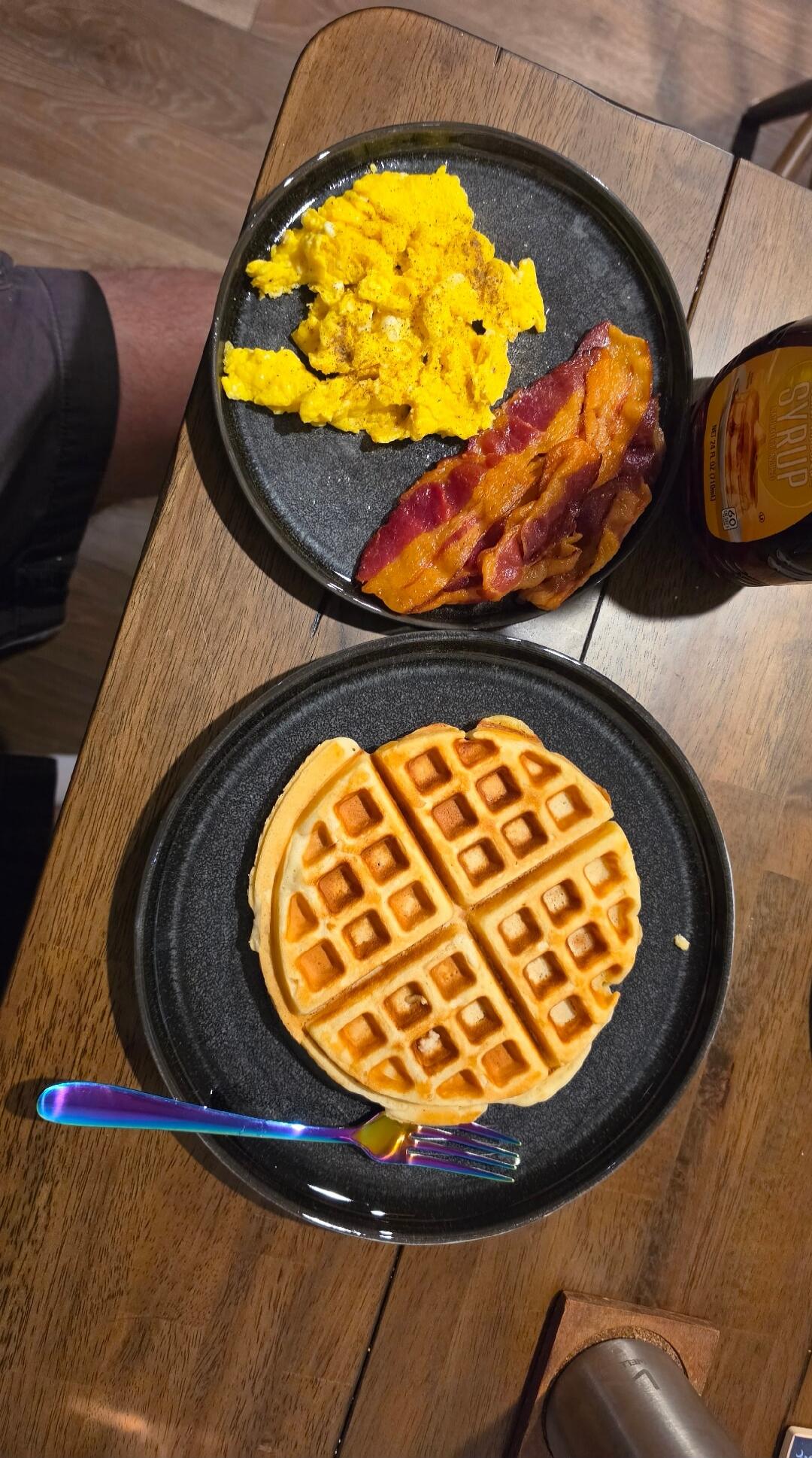 A delicious breakfast spread featuring fluffy scrambled eggs, crispy bacon, and a golden waffle, served with a bottle of syrup and a vibrant iridescent fork.