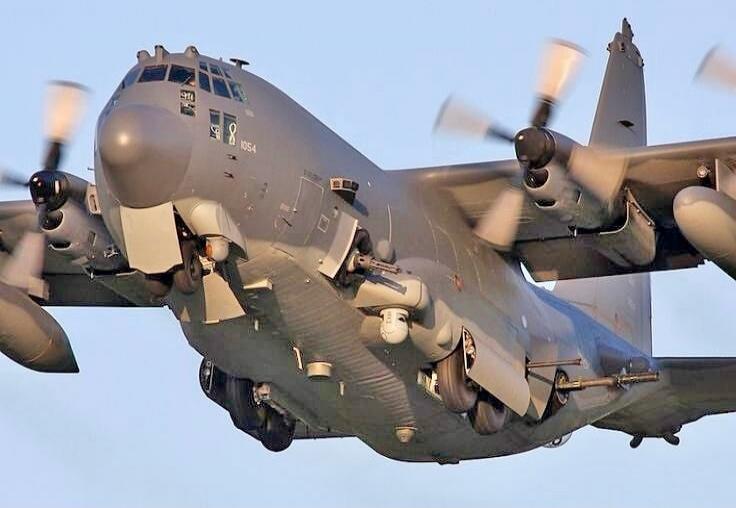 An AC-130 gunship in flight, showing its side-mounted weaponry. The number '8 1054' is visible on the fuselage.