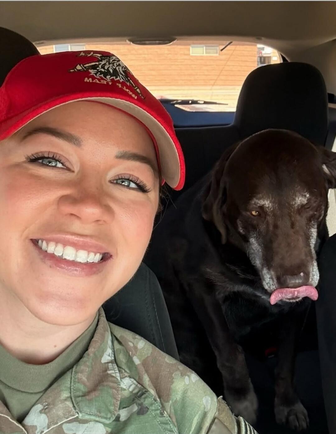 A woman in a military uniform and a red cap is taking a selfie with a brown dog in a car. The woman is smiling, and the dog has its tongue out.