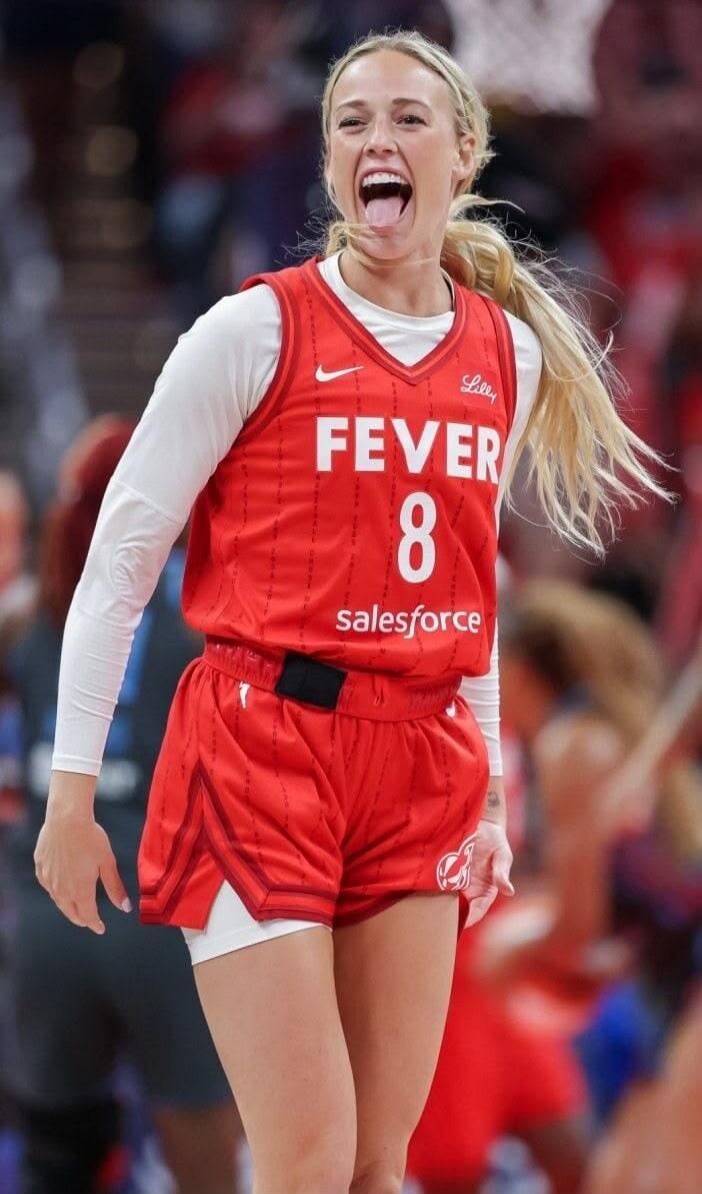 FEVER 8 Lilly salesforce Nike swoosh logo. A female basketball player with blonde hair in a ponytail, wearing a red jersey and white long-sleeve undershirt, making an expressive face with her tongue out during a game.