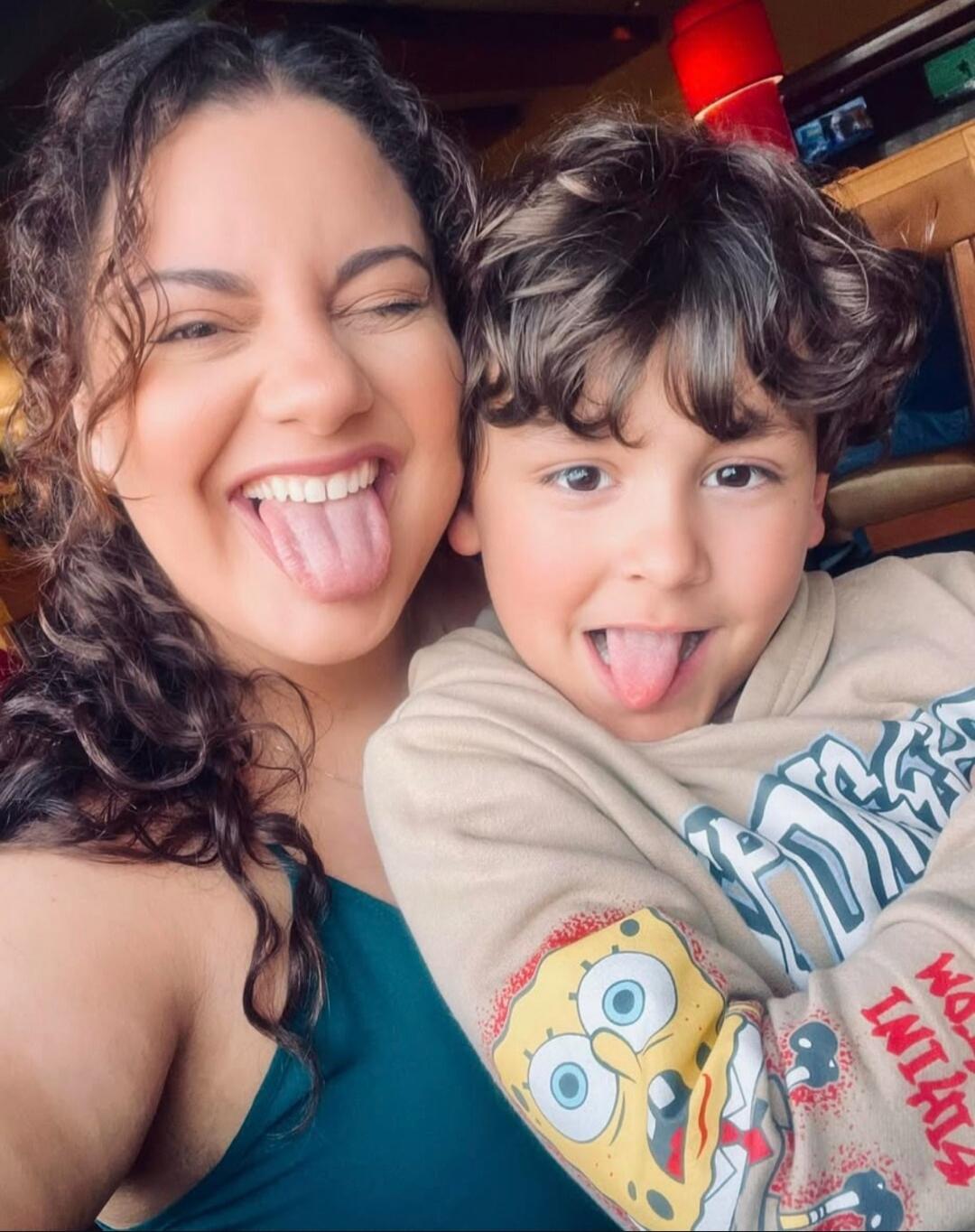 A woman and a young boy, likely mother and son, are making funny faces by sticking out their tongues. The woman is winking with one eye, and the boy is wearing a light-colored hoodie with a Spongebob Squarepants design on the sleeve. They appear happy and playful.