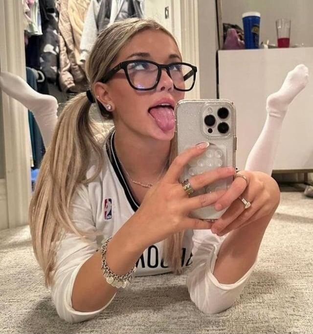 A young woman with blonde pigtails and glasses is lying on the floor, taking a selfie with her phone. She has her tongue sticking out and is looking upwards playfully. Her legs are bent with feet up.